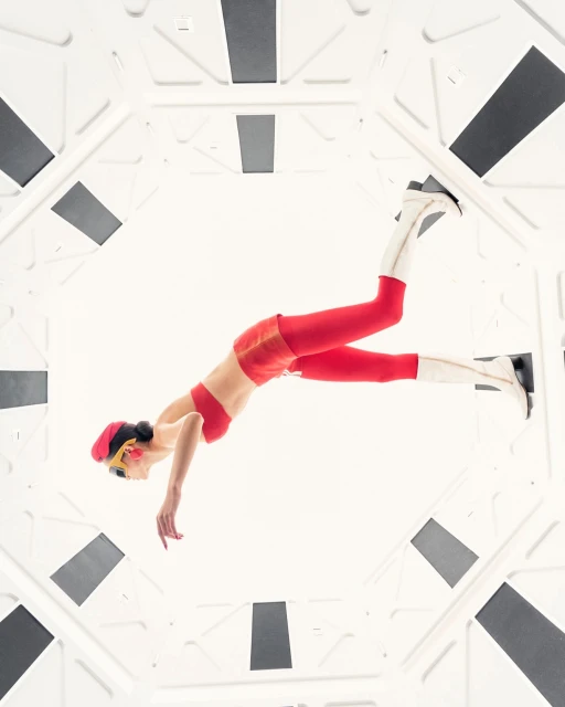 Woman in red sportswear and white boots poses in a futuristic white interior with dark gray panels, shot from a low angle.
