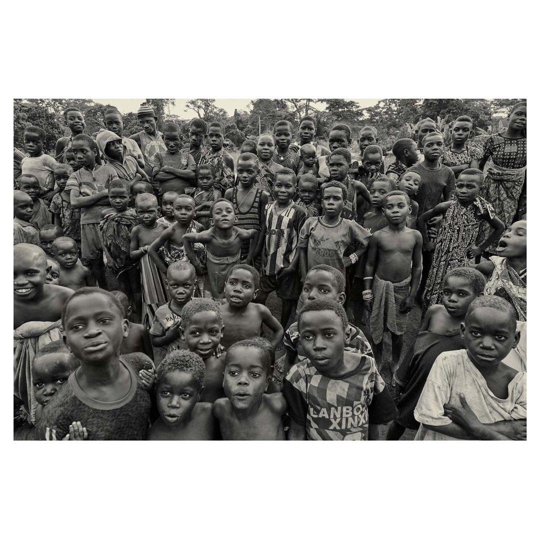 Large group of African children gathered outdoors, mostly shirtless, staring at the camera in a black and white photo.