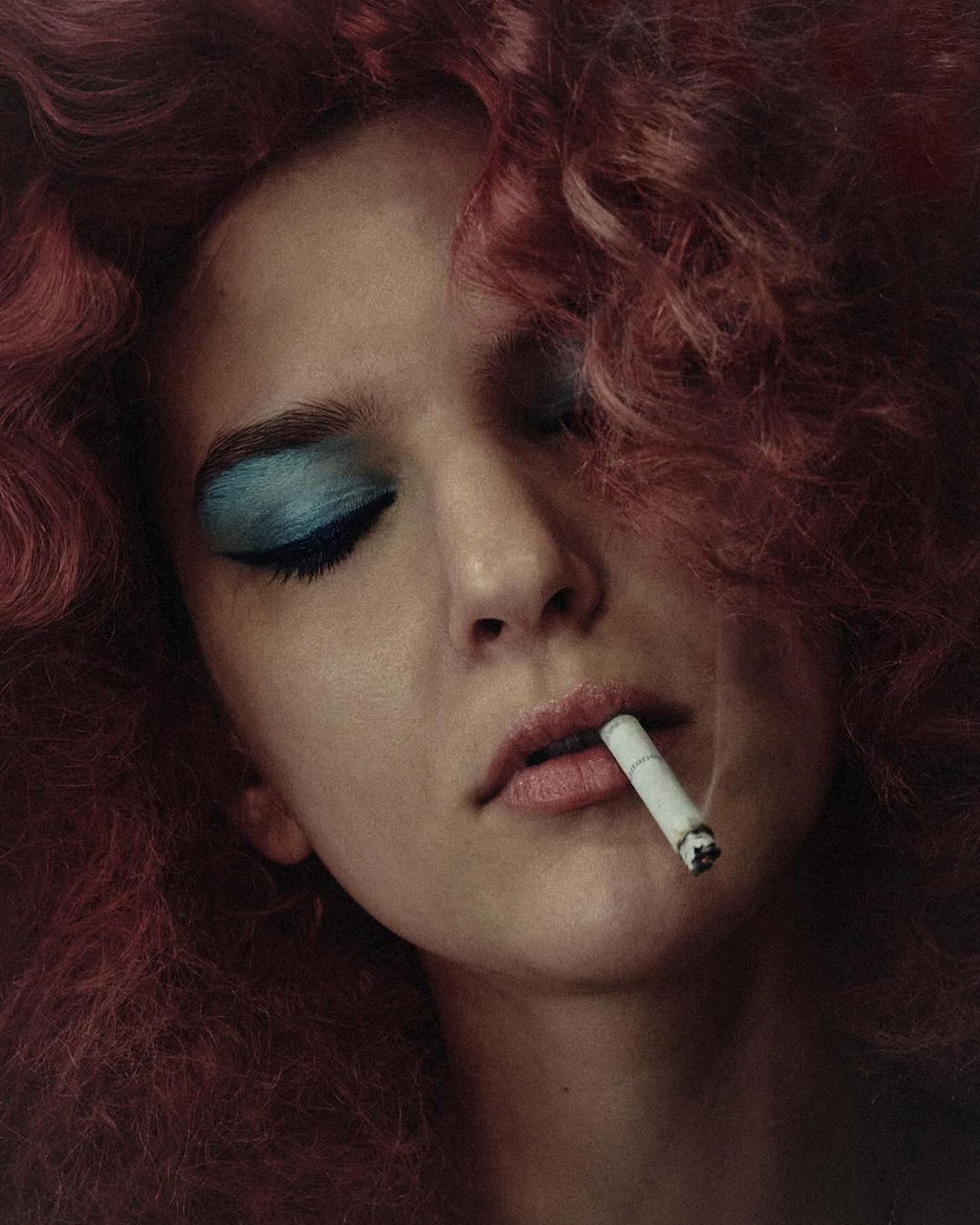 Close-up portrait of a woman with pink curly hair, blue eyeshadow, and cigarette in her mouth, showing beauty and fashion style.