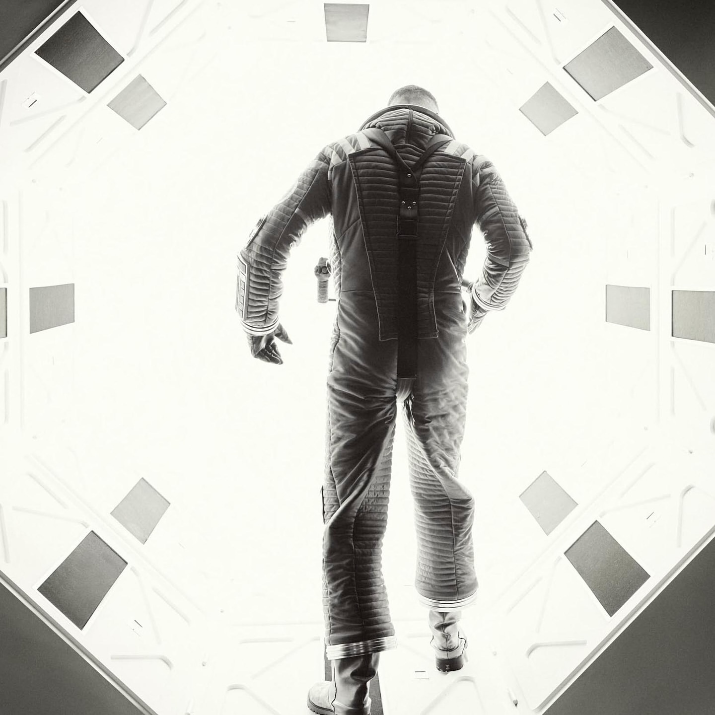 Monochrome image of an astronaut in a space suit walking in a brightly lit white corridor with geometric panels on the sides and ceiling.