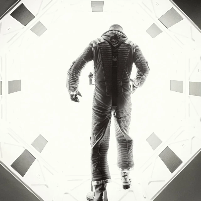 Monochrome image of an astronaut in a space suit walking in a brightly lit white corridor with geometric panels on the sides and ceiling.