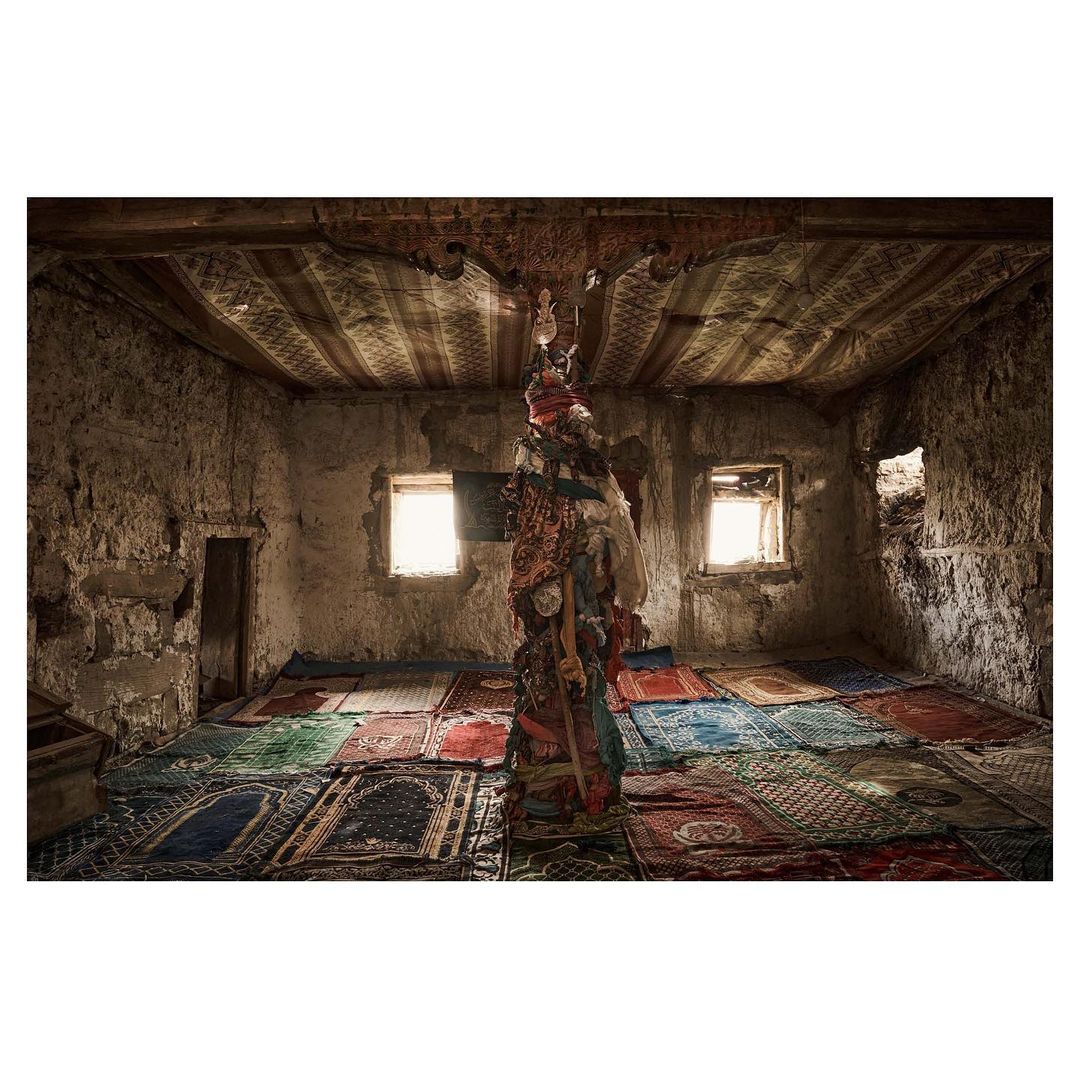 Interior of weathered room with ornate rugs, dilapidated walls, and a column draped with fabric, illuminated by natural light.