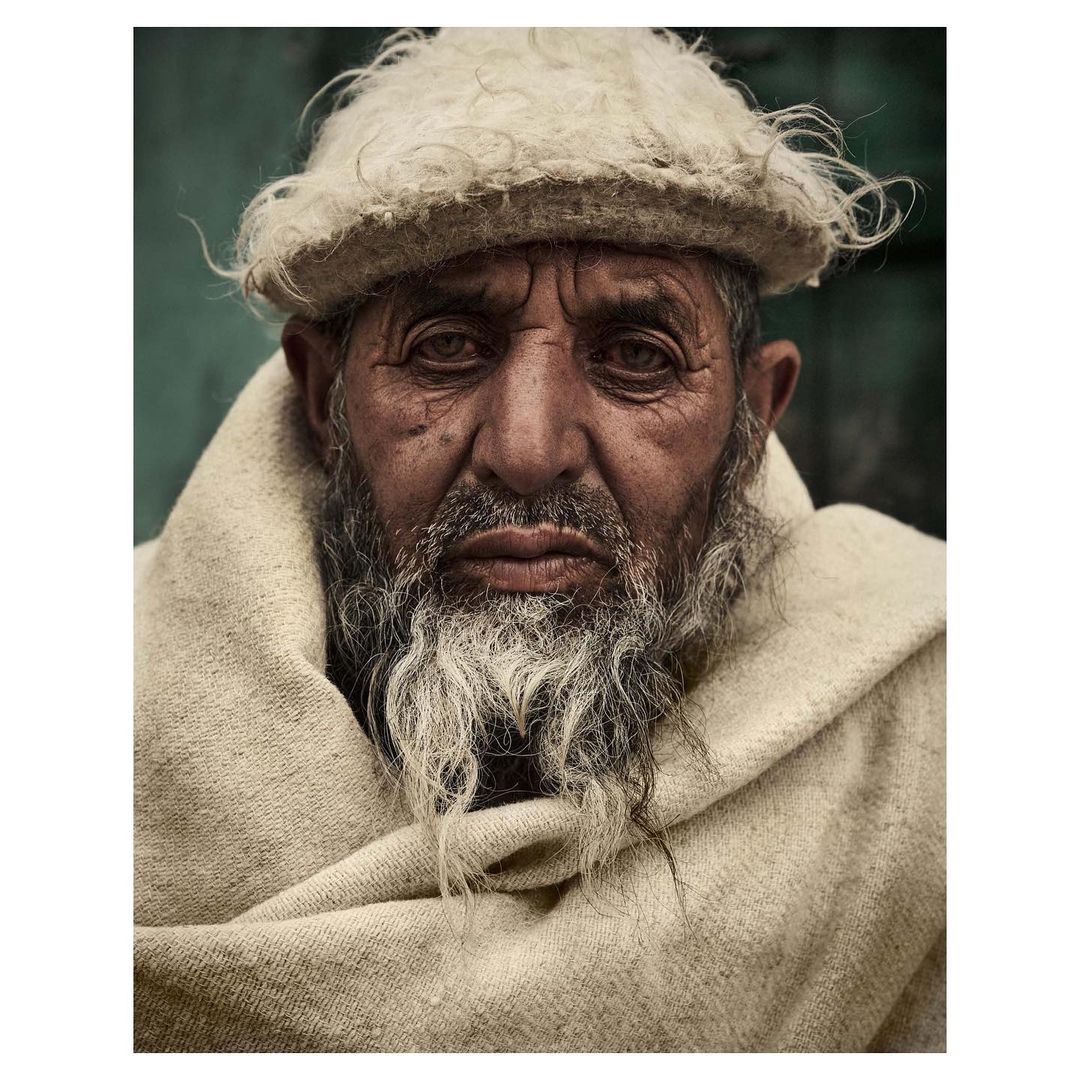 Close-up portrait of an elderly Middle Eastern man with a white beard wearing a traditional white wool hat and shawl, looking at the camera.