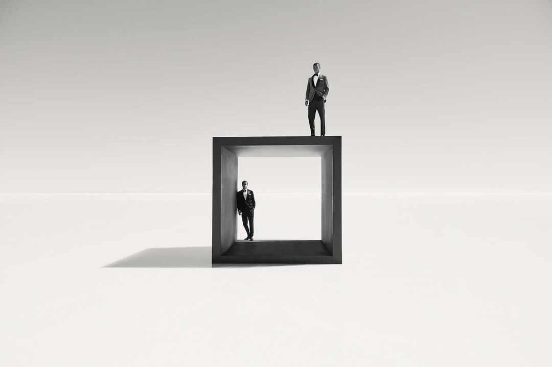 Monochrome photograph of two adult males in formal wear, one standing on top of a black cube and the other standing within it.