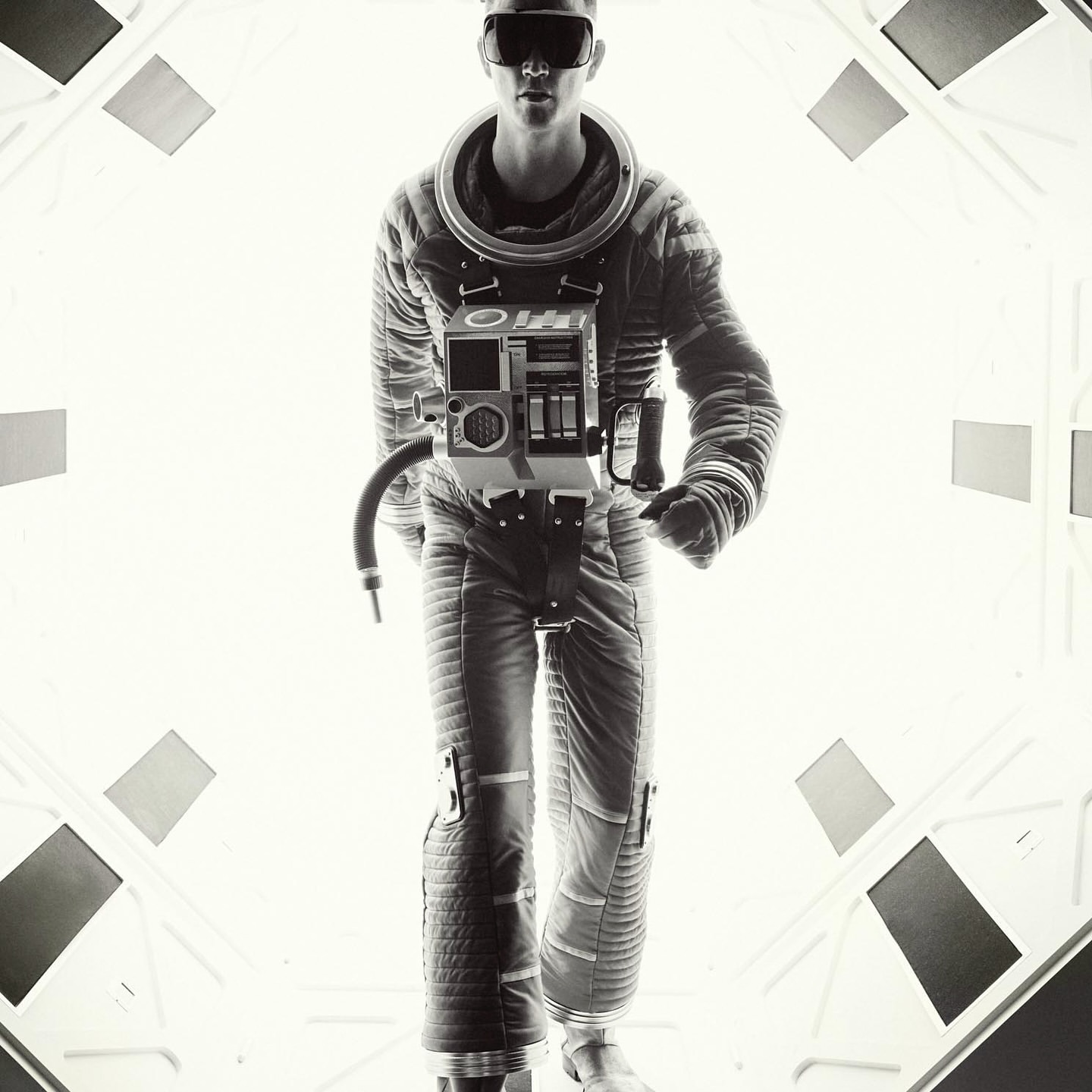 Monochrome, full shot of a man in a spacesuit walking in a hallway with geometric patterns and bright, diffused lighting. 