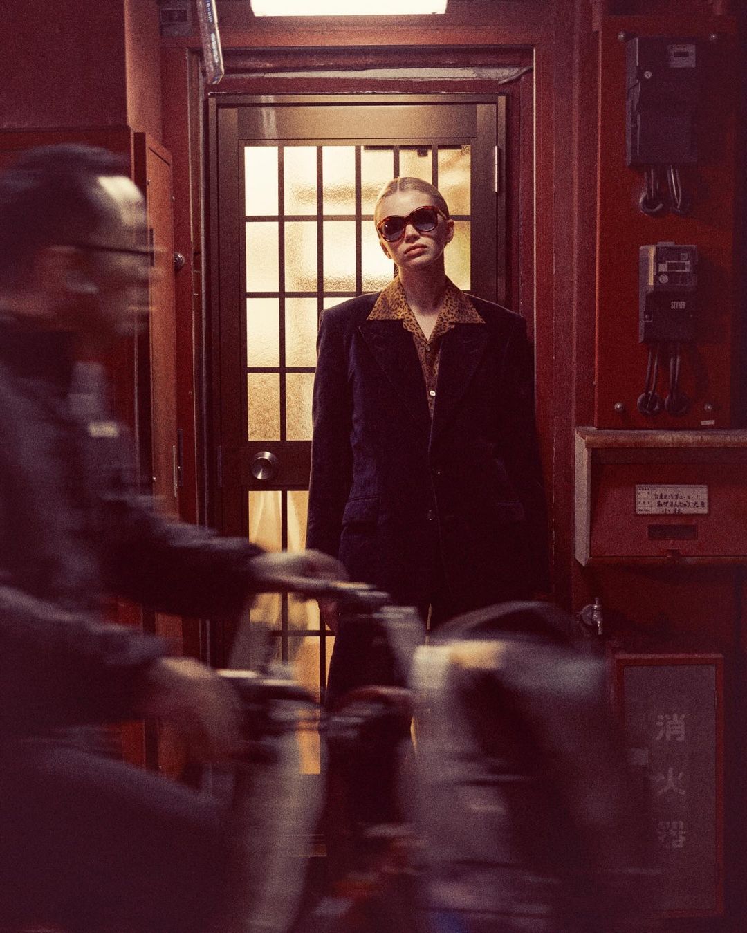 Stylish blonde woman in sunglasses and blazer stands in a doorway, with a blurred person in the foreground adding motion.