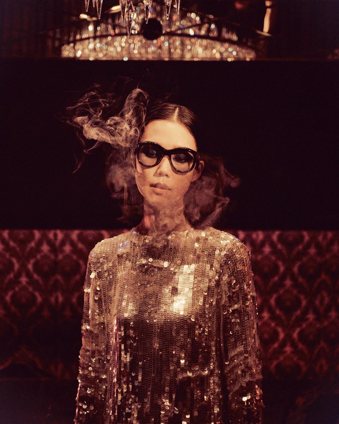 Woman with black-framed glasses wearing a gold sequin dress, smoke in air, against a dark background with chandelier.