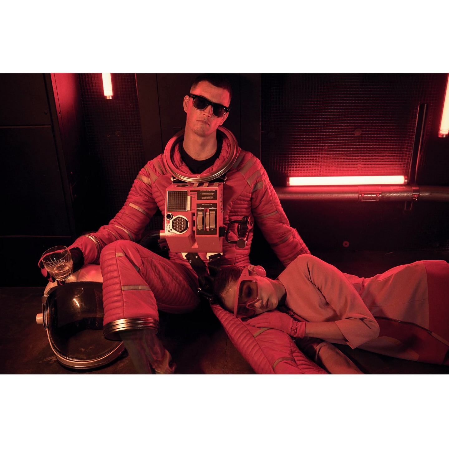 A male figure in a red astronaut suit with sunglasses sits indoors beside a woman with a glass; red neon tubes in the background.