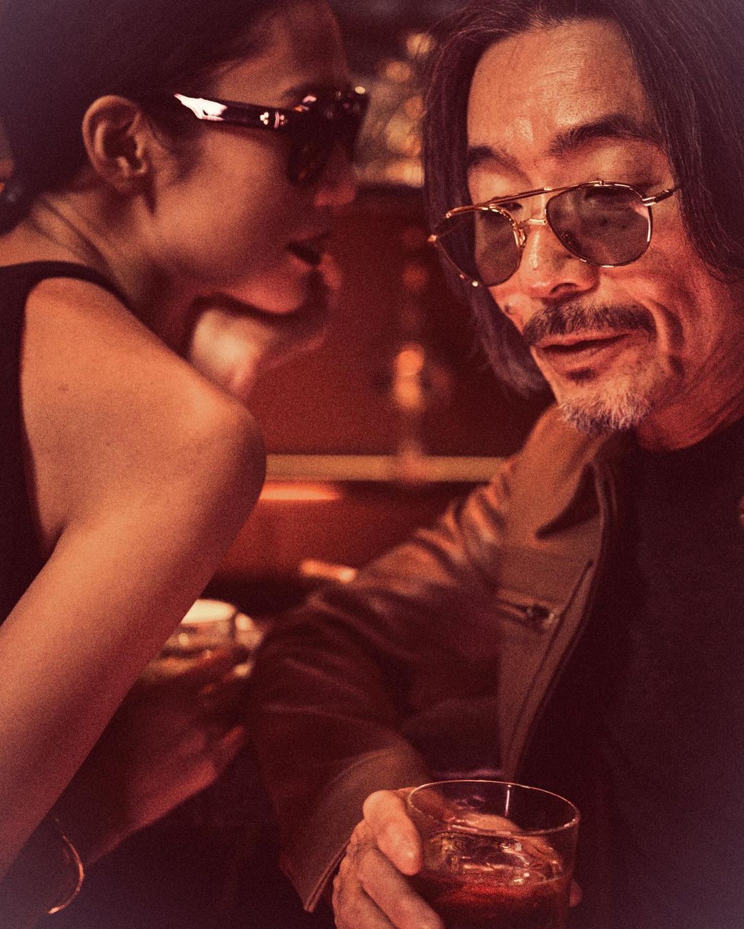 Man with sunglasses holds a drink while a woman leans in, both in semi-formal attire, at an indoor bar setting with warm, soft lighting.