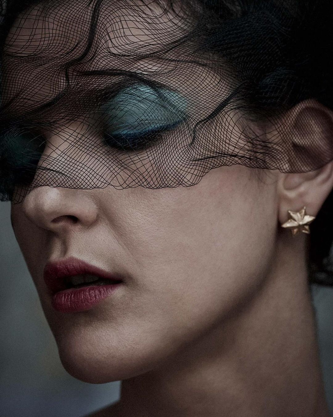 Close-up portrait of a woman with dramatic makeup and black netting obscuring the upper half of her face, wearing star-shaped earrings.
