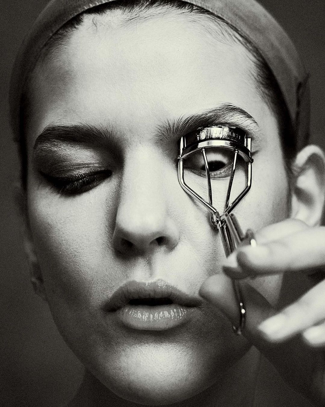Close-up black and white portrait of a woman using an eyelash curler with one eye closed and the other open, wearing a headband.