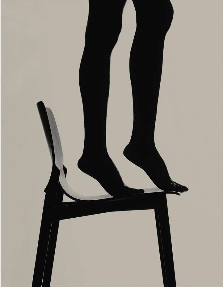 Silhouette of legs and feet standing on a black chair against a light background, creating a minimalist and artistic composition.