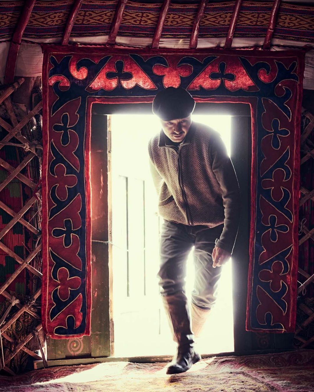 An adult male standing in the doorway of a yurt, wearing a jacket and jeans, with an ornate red and black fabric door frame.