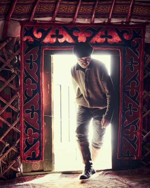 An adult male standing in the doorway of a yurt, wearing a jacket and jeans, with an ornate red and black fabric door frame.