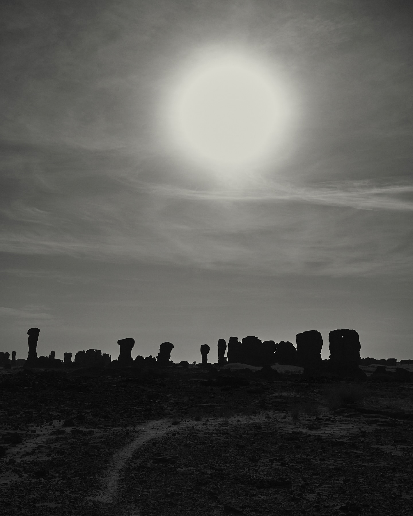 Monochrome landscape with rock formations silhouetted against the sky and sun, creating a stark contrast in a sparse, outdoor setting.