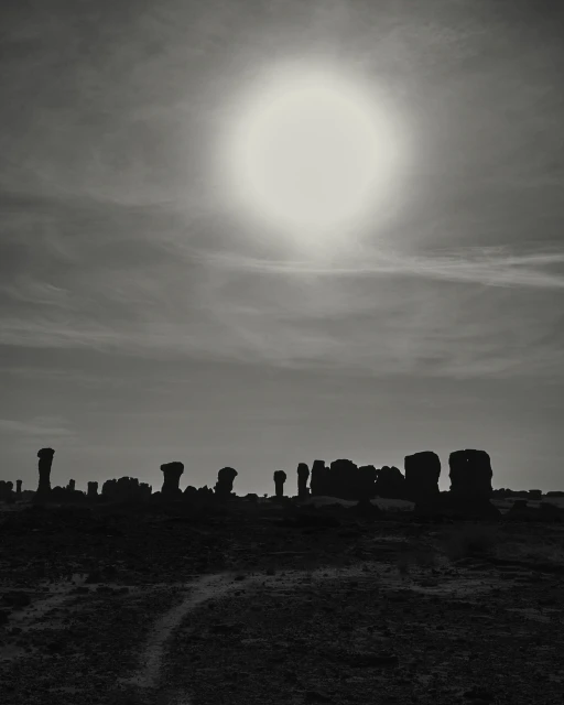 Monochrome landscape with rock formations silhouetted against the sky and sun, creating a stark contrast in a sparse, outdoor setting.
