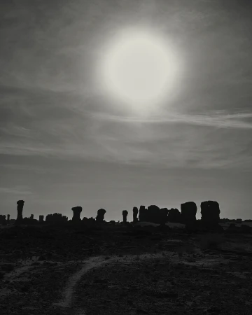 Monochrome landscape with rock formations silhouetted against the sky and sun, creating a stark contrast in a sparse, outdoor setting.