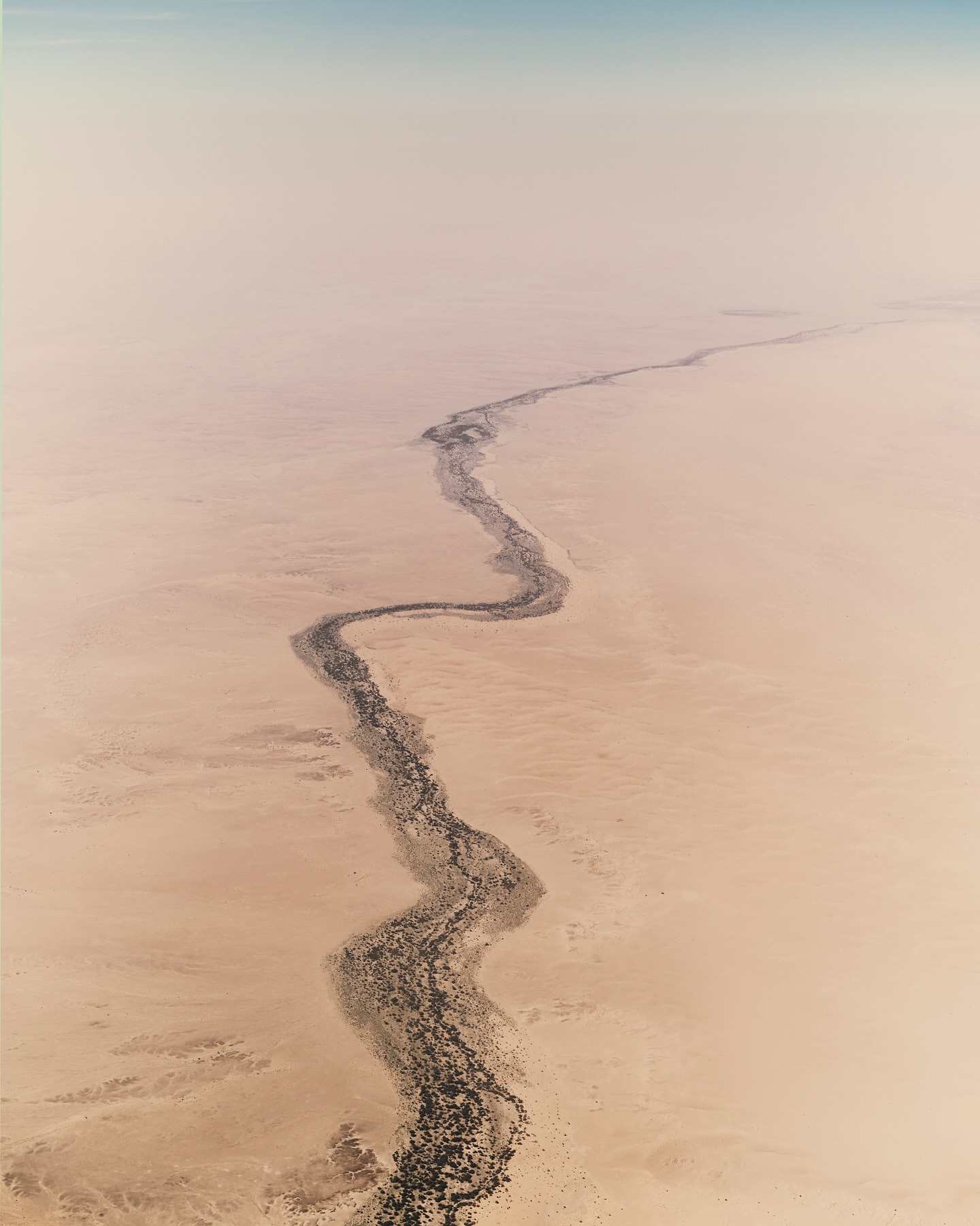 Aerial view of a meandering riverbed cutting through a vast, tan desert landscape under a hazy, light-blue sky; geological formations.