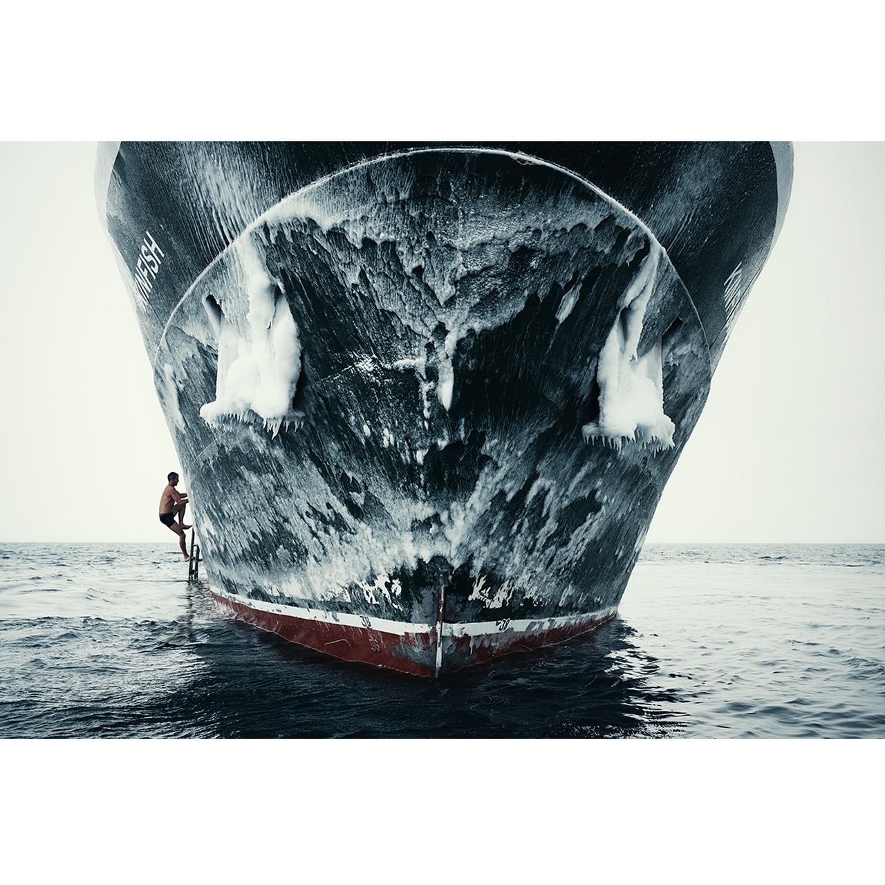 A man climbs a ladder onto a large, weathered cargo ship in the ocean, captured in a dramatic wide shot, under an overcast sky.