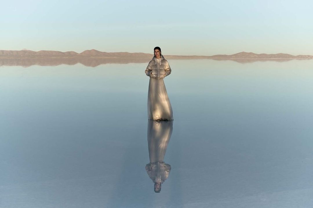 Adult female standing in reflective water wearing a silver puffer coat dress with distant mountains and a pastel blue sky.