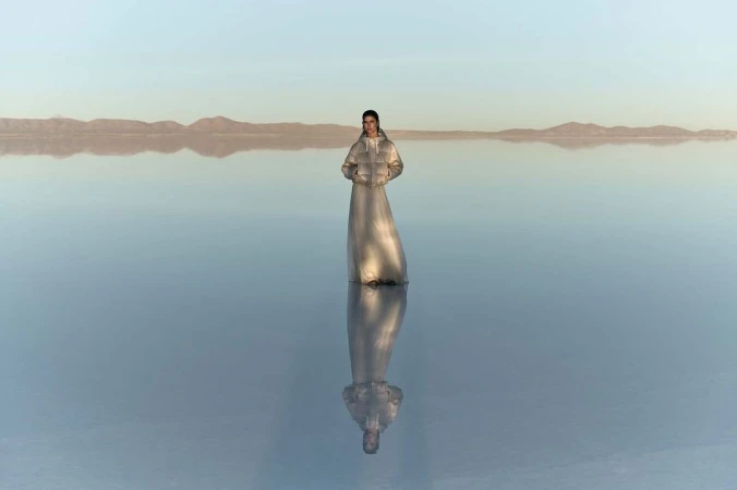 Adult female standing in reflective water wearing a silver puffer coat dress with distant mountains and a pastel blue sky.