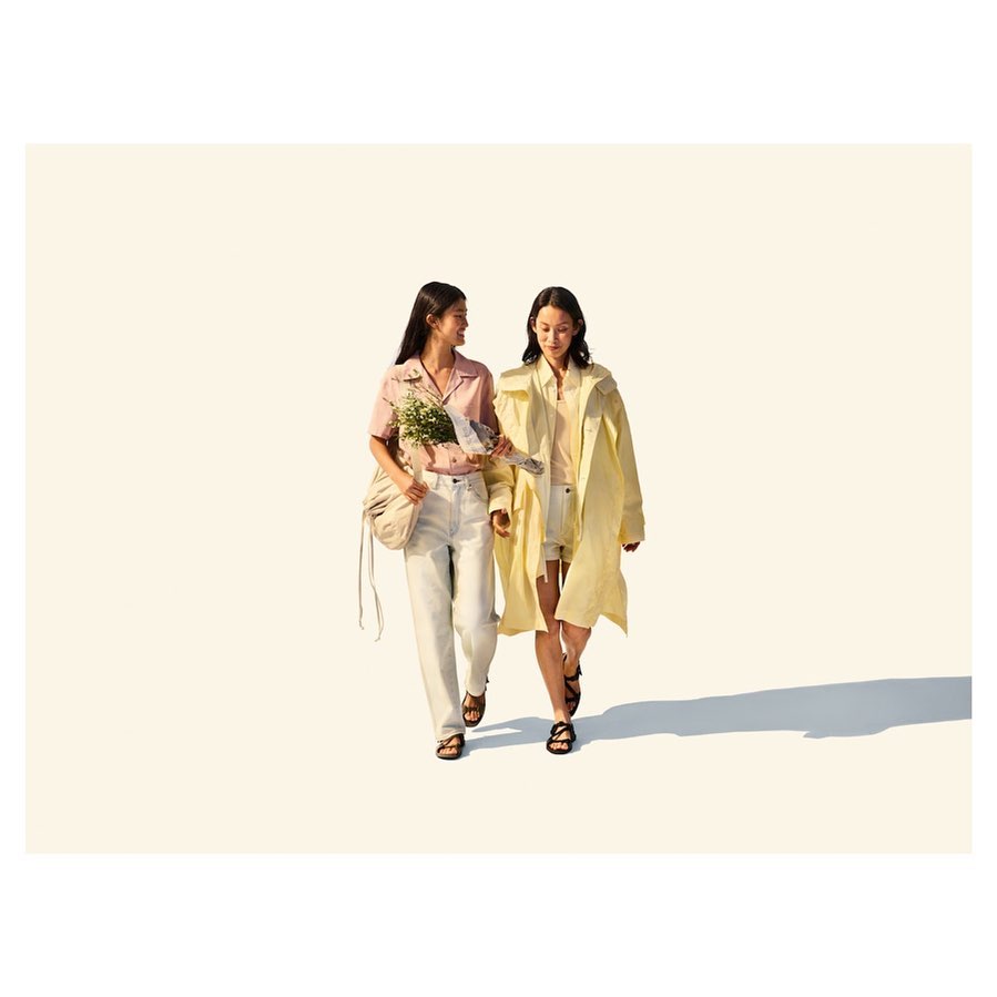 Two women walking in a studio, one carries flowers, both wear sandals. Neutral background with soft lighting and shadows. 