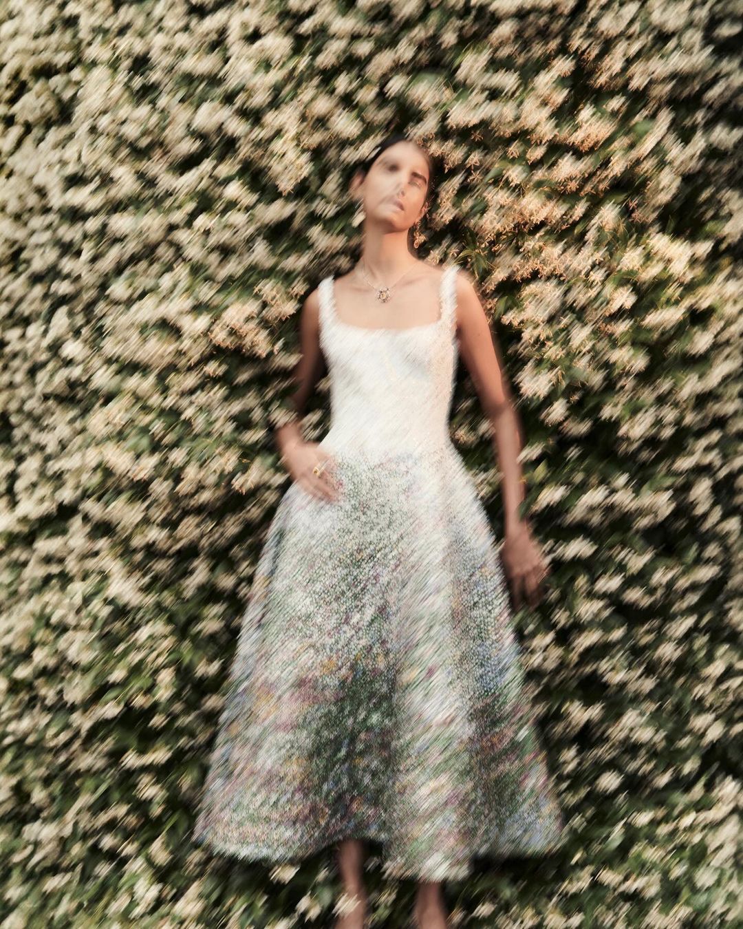 Woman in flowing floral dress stands in front of flowering white vine wall. Light-skinned, elegant pose, jewelry, neutral expression.