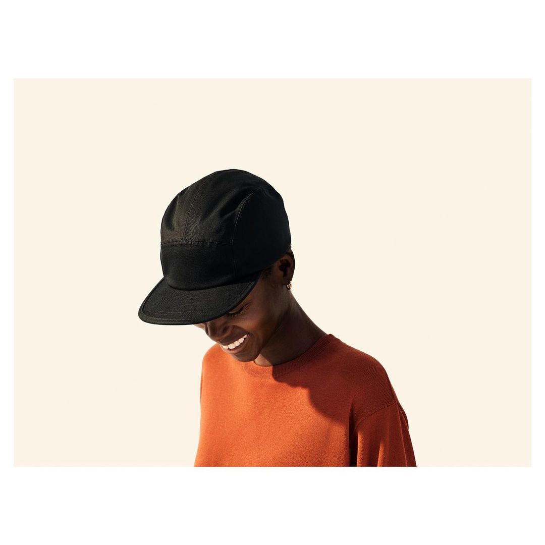 Smiling adult wearing a black cap and an orange shirt against a neutral backdrop. The person is lit by a soft light source.
