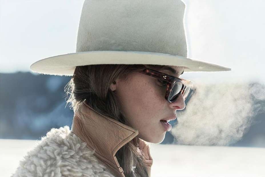 Portrait of a woman with hat and sunglasses exhaling vapor into cold outdoor air, wearing a tan coat with fur collar, against a neutral background.
