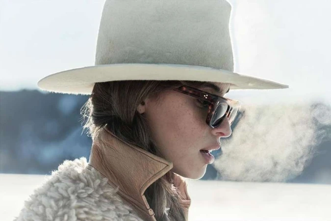 Portrait of a woman with hat and sunglasses exhaling vapor into cold outdoor air, wearing a tan coat with fur collar, against a neutral background.