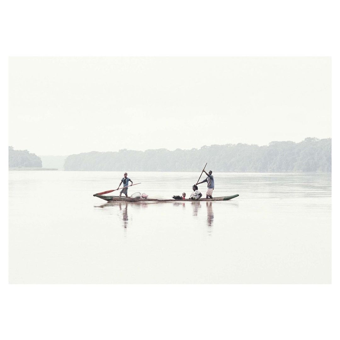 Four people in a narrow wooden canoe on a wide river, using poles to navigate, with lush green vegetation on the distant shoreline.