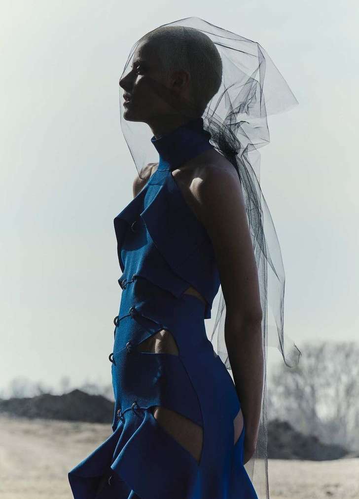 A woman with short hair and a blue dress poses outdoors with a black veil. Fashion photography showcases modern haute couture.