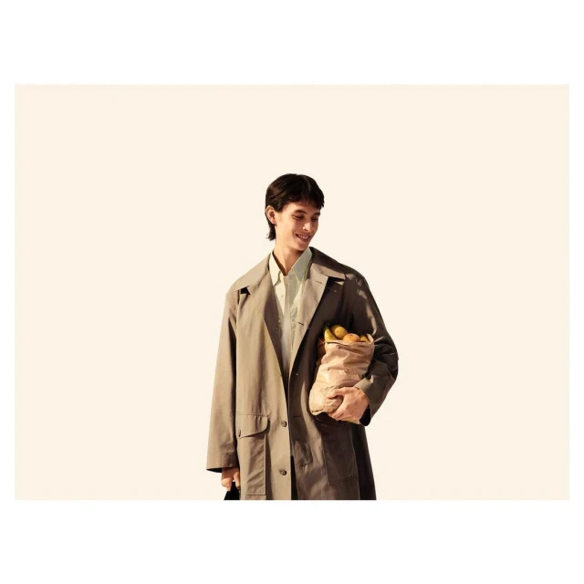 Adult male in beige trench coat smiles holding grocery bag outdoors with bright lighting and neutral background in lifestyle fashion.