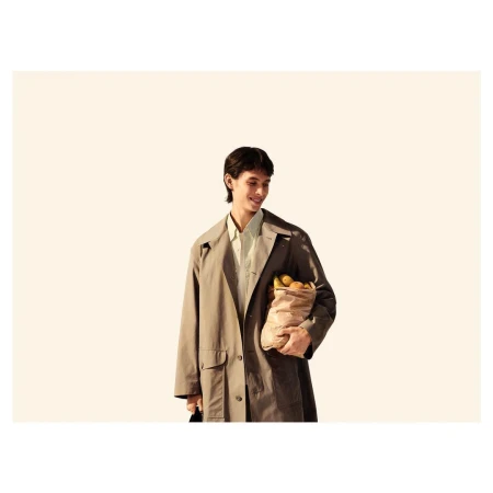 Adult male in beige trench coat smiles holding grocery bag outdoors with bright lighting and neutral background in lifestyle fashion.