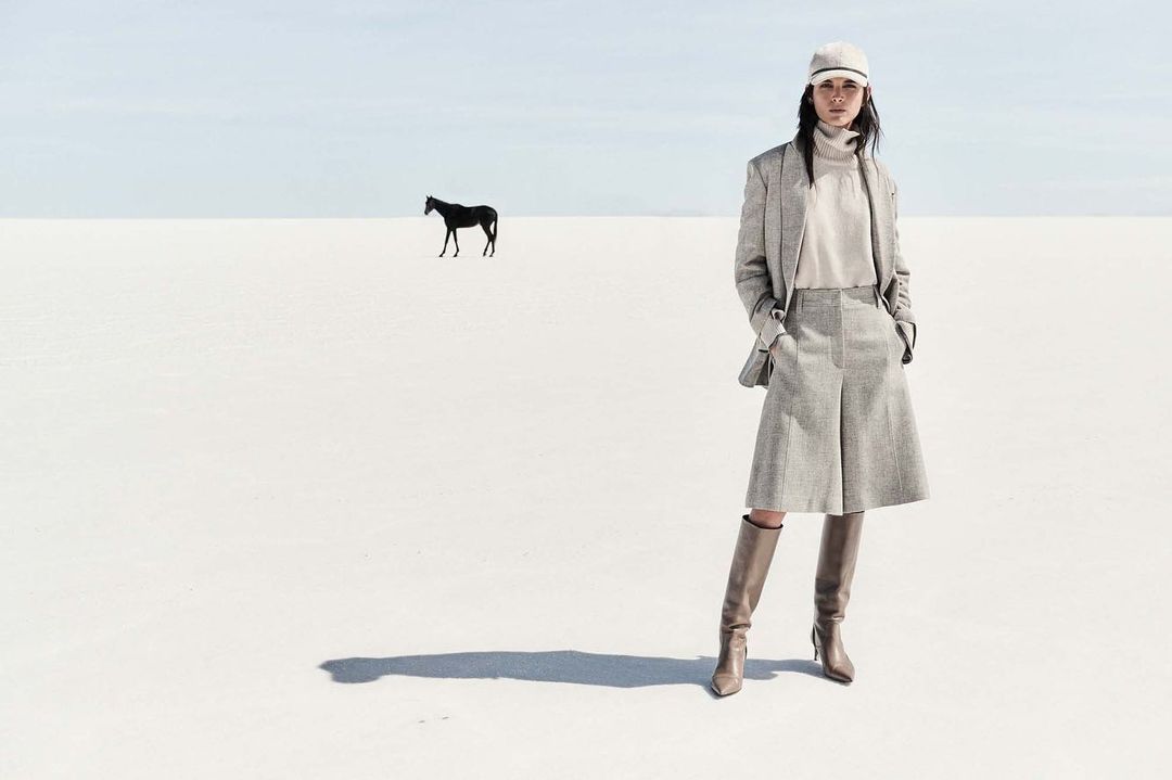 Woman in tweed outfit and tall boots poses in white landscape, with black horse in the distance under a light blue sky.