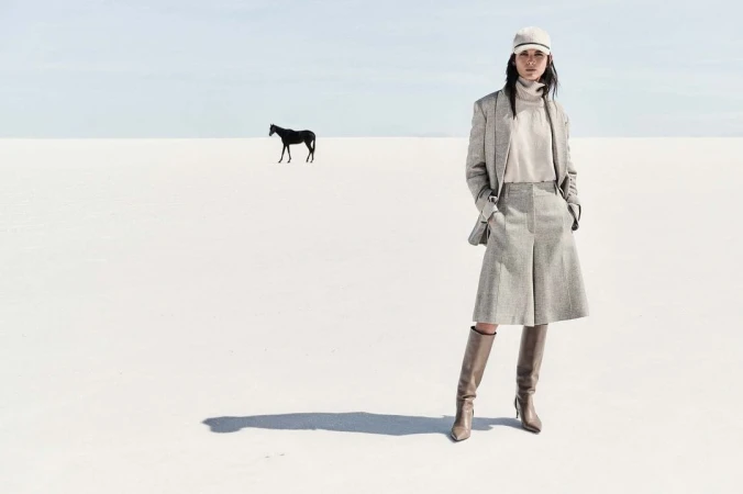 Woman in tweed outfit and tall boots poses in white landscape, with black horse in the distance under a light blue sky.