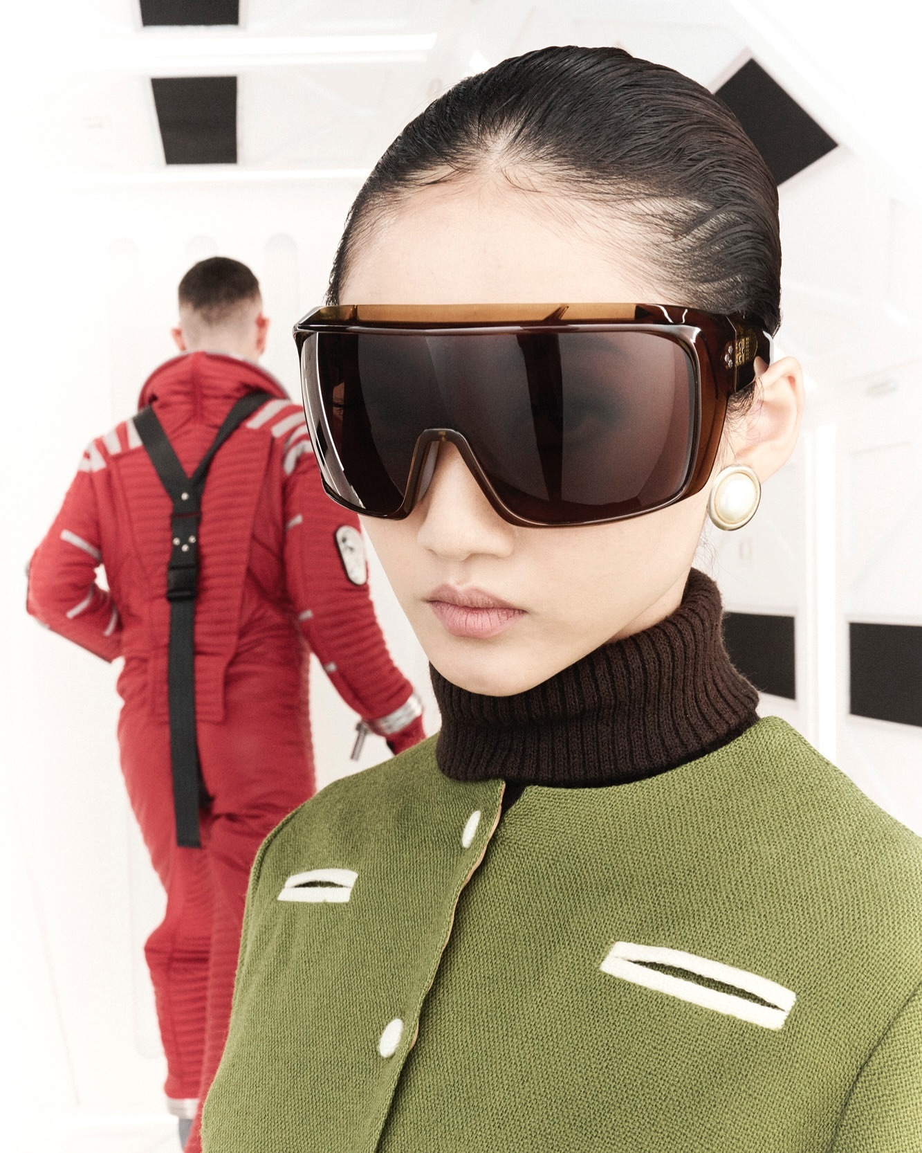 A stylish woman in sunglasses and a green jacket stands in a futuristic setting, with a male astronaut in red jumpsuit behind her.