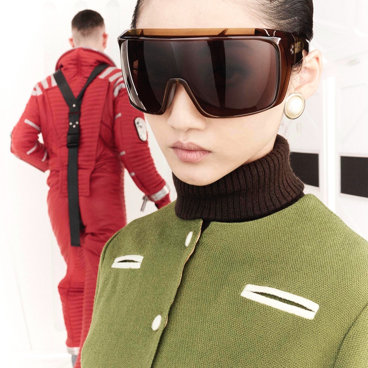 Fashion shot of a woman wearing oversized sunglasses and a green jacket, with a man in a red jumpsuit in the background, studio setting.
