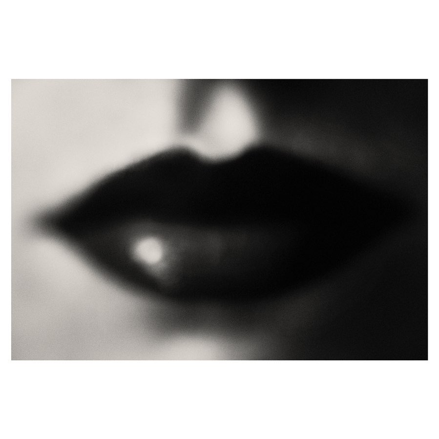 Close-up of blurred, dark red lips showing a single point of reflected light, set against a soft, out-of-focus light grey background.