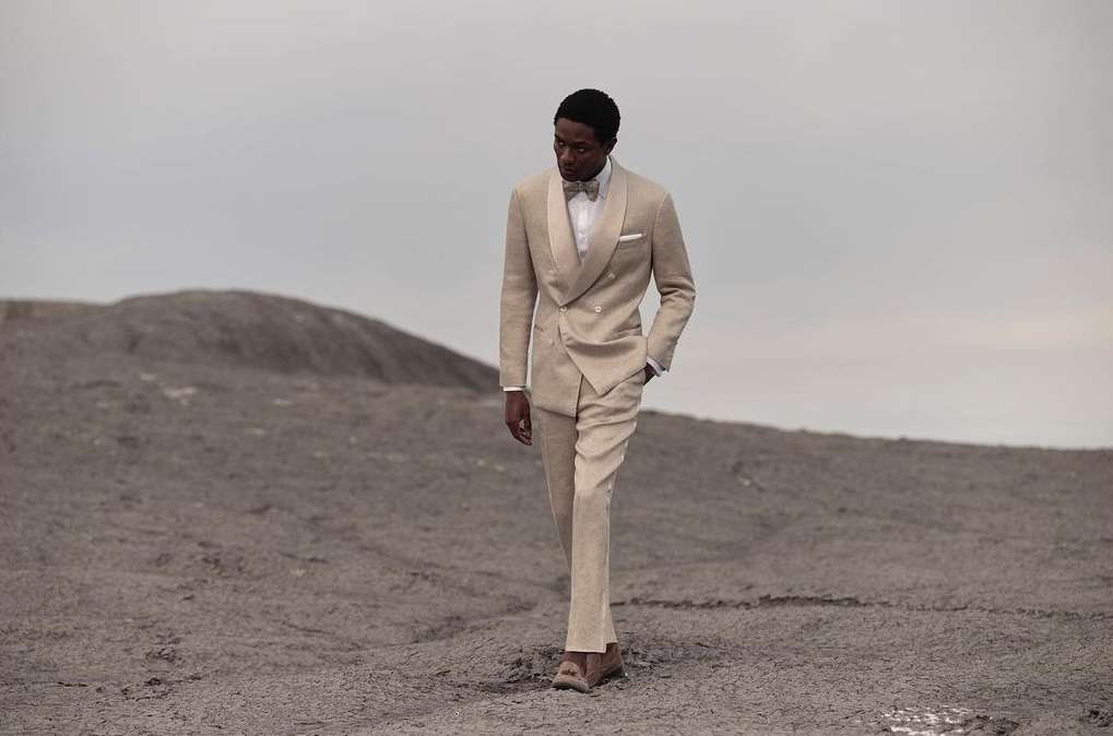 An adult black man in a cream double-breasted suit walks across a gray desert landscape under a cloudy sky in an outdoor fashion editorial.