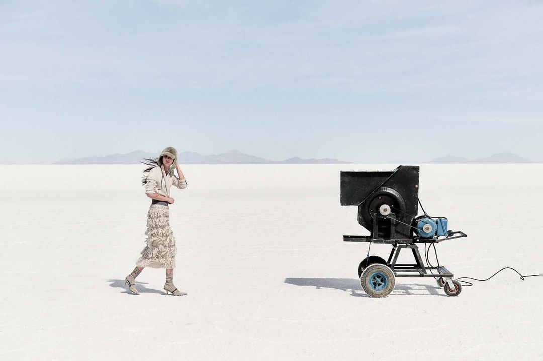 A woman in a fringed skirt walks across a white landscape toward a large black industrial machine on wheels under a clear blue sky.
