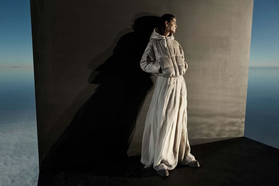 An adult female fashion model poses in a hooded jacket and long skirt against a wall in an abstract outdoor water-sky setting.