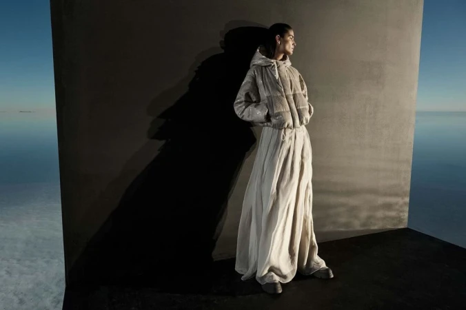An adult female fashion model poses in a hooded jacket and long skirt against a wall in an abstract outdoor water-sky setting.