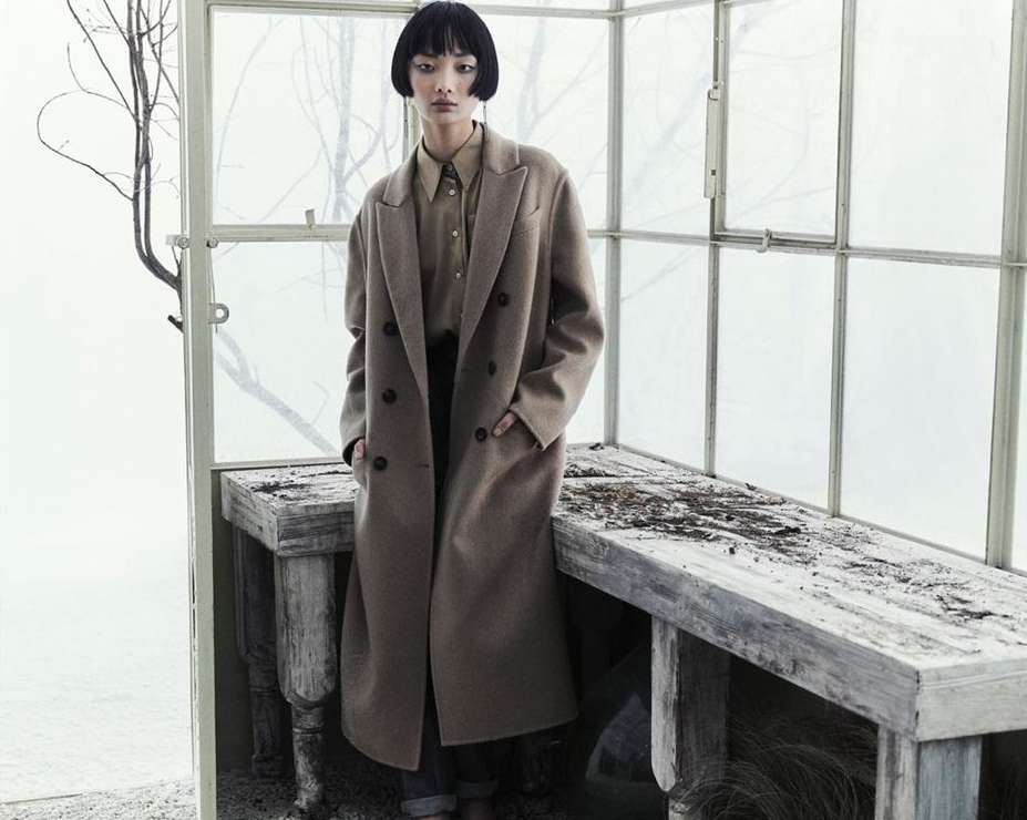 A stylish Asian woman poses indoors in a long brown coat and collared shirt. The setting features textured walls and wood accents.
