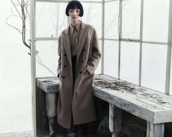 A stylish Asian woman poses indoors in a long brown coat and collared shirt. The setting features textured walls and wood accents.