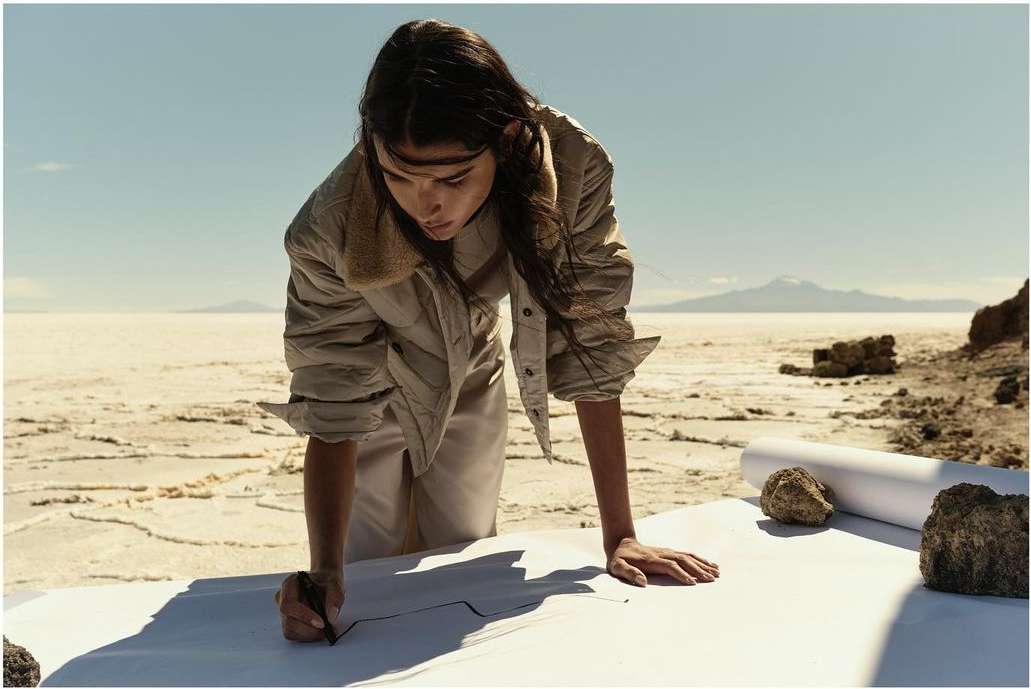 A woman in a jacket drawing on a large sheet of paper outdoors in a desert-like environment, with mountains visible in the distance under a clear sky.