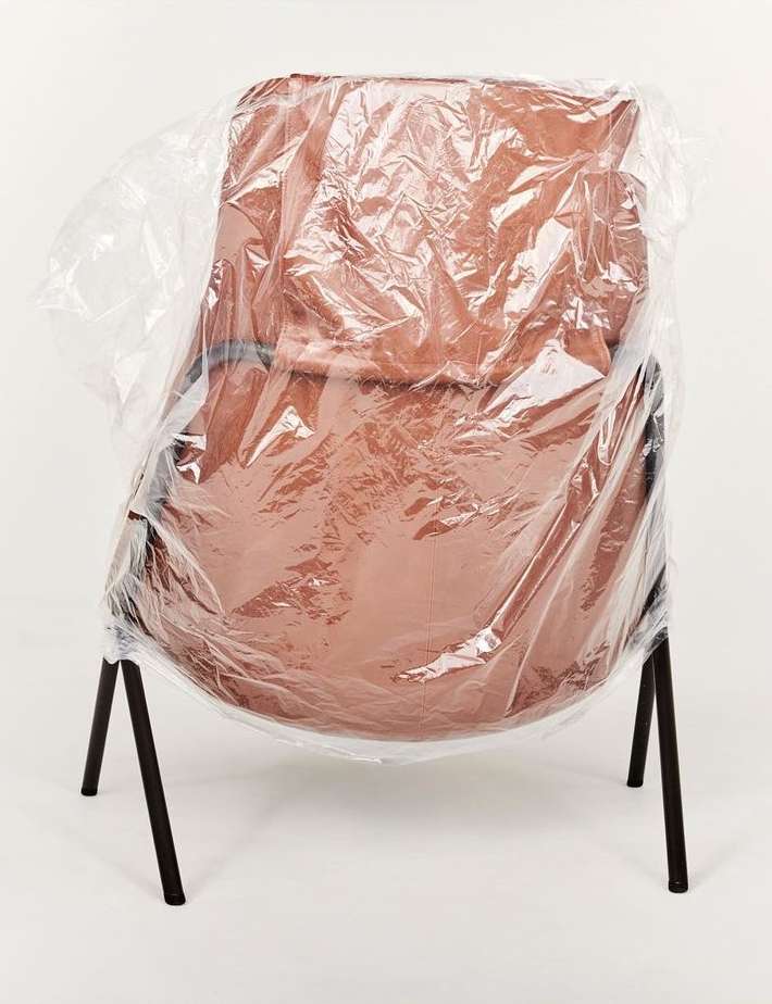 Upholstered chair is covered in clear plastic on a white background indoors, showcasing furniture protection and minimalist presentation.