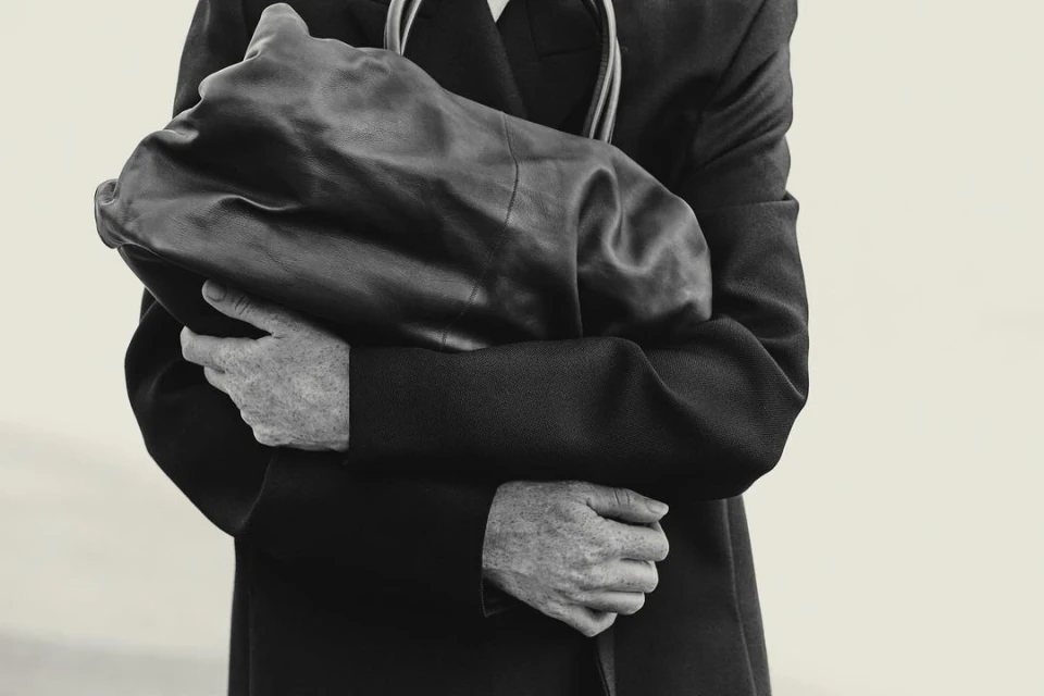 Monochrome portrait of an adult holding a dark leather-like object in front of their chest, wearing a black coat against an off-white backdrop.