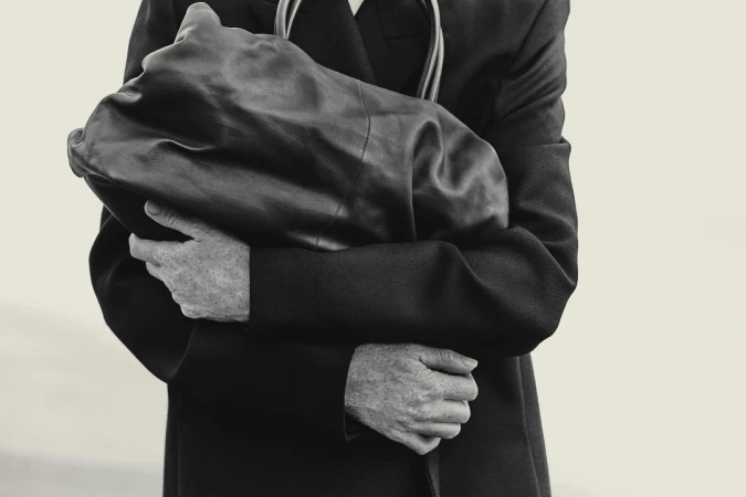 Monochrome portrait of an adult holding a dark leather-like object in front of their chest, wearing a black coat against an off-white backdrop.