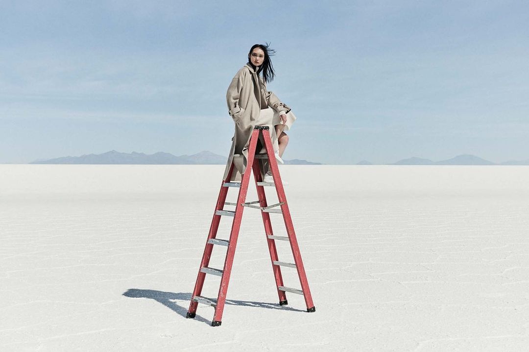 A woman in a beige coat sits on a red ladder in a vast, light desert landscape under a blue sky with distant mountain range.